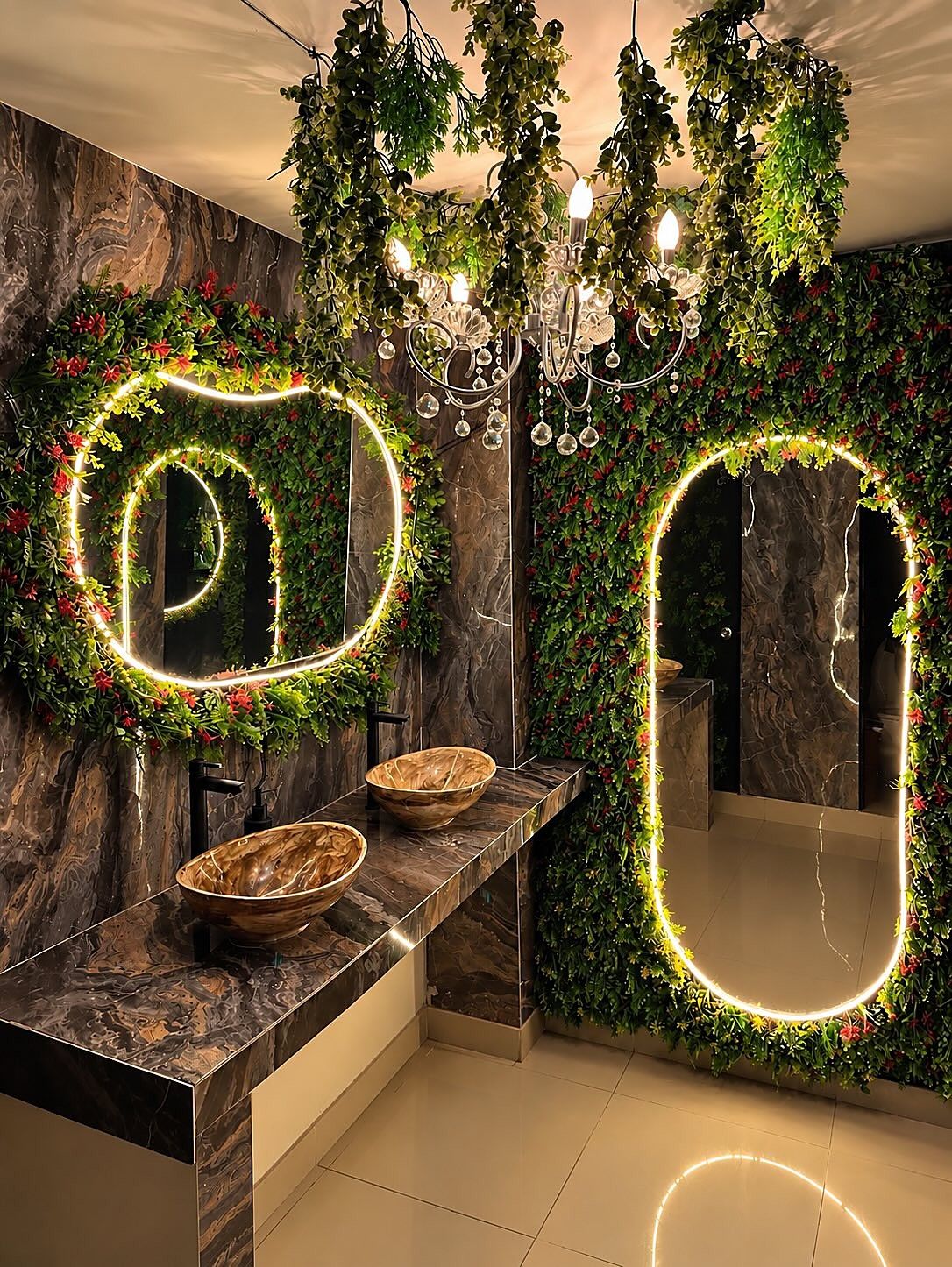 Espejos con aro de luz sobre una pared de plantas, lavamanos de piedra y una lámpara de cristal, en Selva Negra