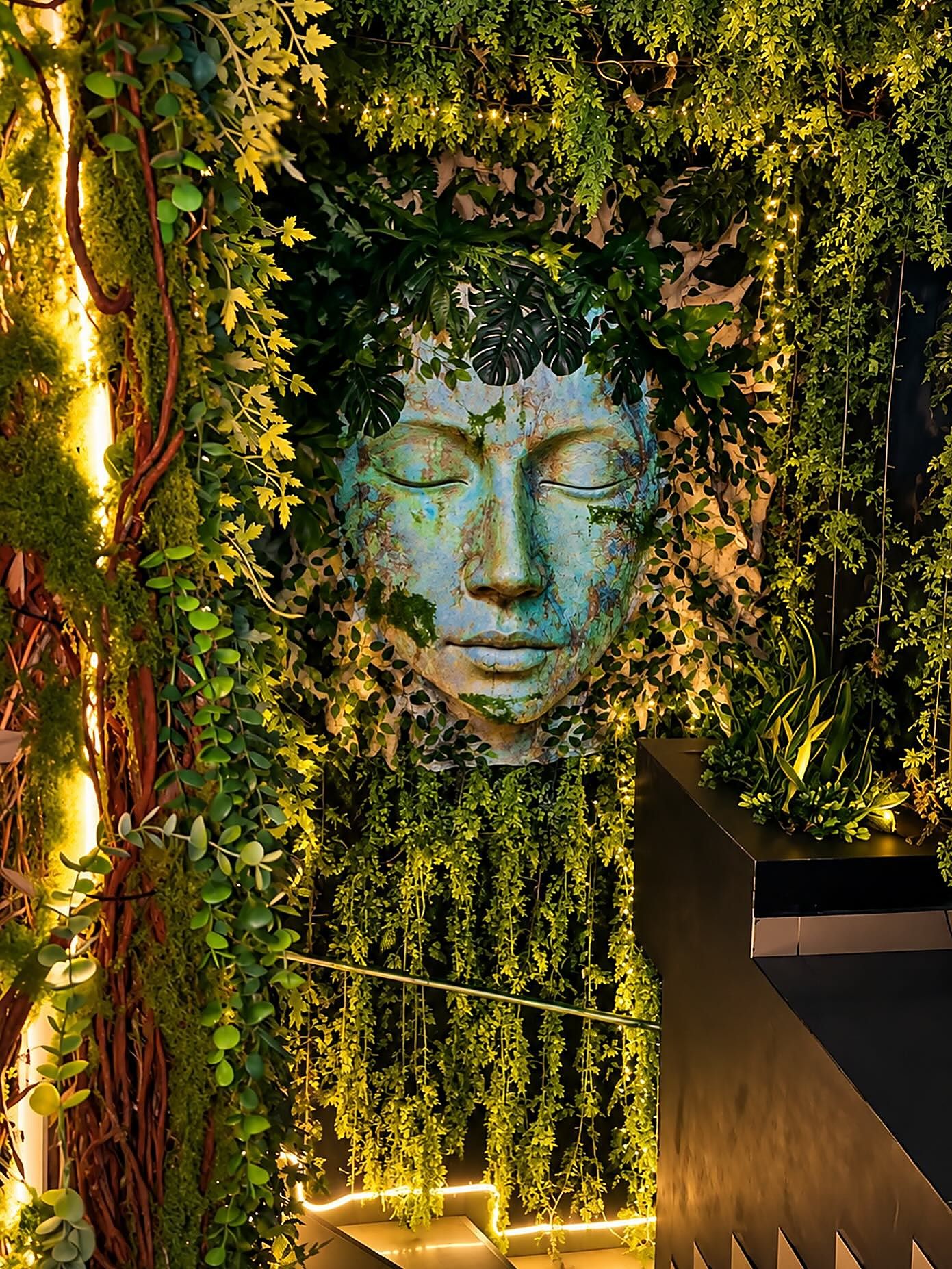 Rostro de piedra turquesa con los ojos cerrados, rodeado de hojas y luces tibias, en una pared de Selva Negra
